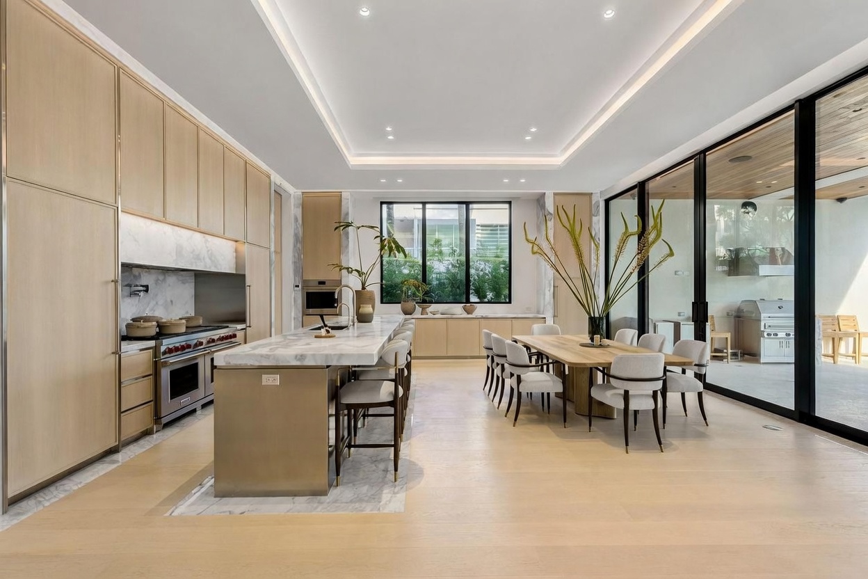 This expansive, modern kitchen and dining area features light wood cabinetry, a large stone-topped island with integrated seating, and a matching dining table. The space is defined by its clean lines, recessed ceiling lighting, and floor-to-ceiling glass walls that provide a seamless transition to the outdoor patio. The neutral color palette and minimalist aesthetic create an airy, sophisticated atmosphere suitable for high-end living.