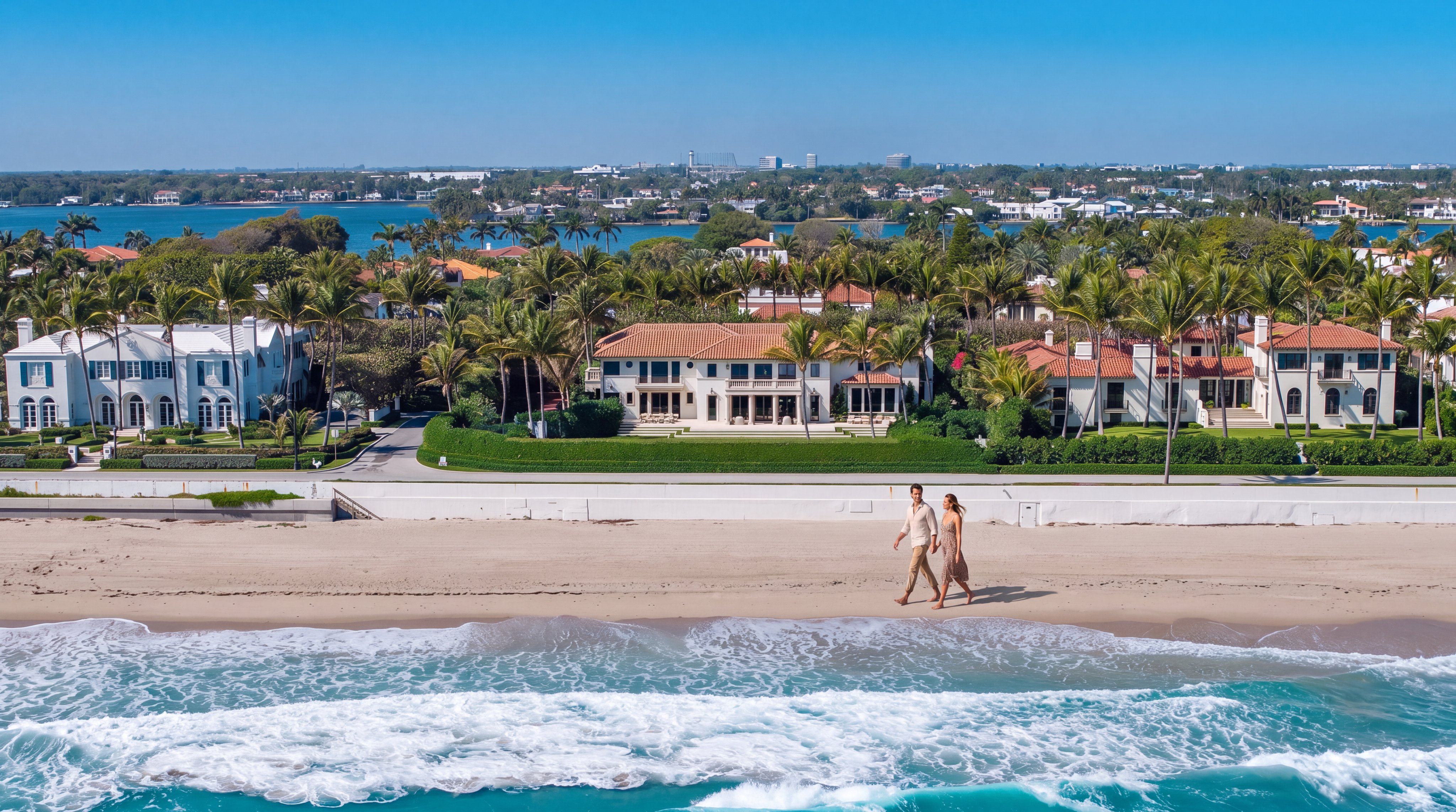 This stunning aerial view captures a row of luxurious, Mediterranean-inspired beachfront estates along a pristine sandy coastline. A couple is seen walking along the beach, highlighting the direct waterfront access and the serene, upscale residential environment. The properties feature manicured lawns, red-tiled roofs, and lush landscaping, set against the backdrop of a vibrant blue ocean and clear sky.