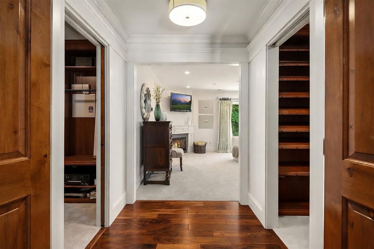 This perspective captures a transition space featuring rich, dark wood flooring that leads from a hallway into a carpeted living area. The hallway is flanked by two built-in storage areas with wooden shelving, while the living room beyond showcases a fireplace with a mantel, a wall-mounted television, and a large window draped in light-colored curtains. The overall aesthetic is traditional and well-maintained, with crisp white trim and crown molding providing a clean, cohesive look.