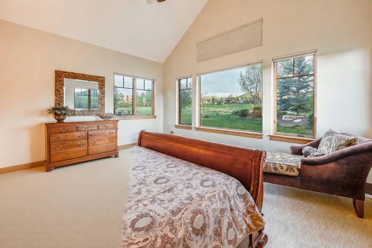 This spacious primary bedroom features a vaulted ceiling and a large, ornate wooden bed frame as the central focal point. A matching wooden dresser with a decorative mirror sits against the wall, while a plush chaise lounge is positioned near the expansive windows that offer a scenic view of the surrounding landscape. The room is decorated in warm, neutral tones, creating a comfortable and inviting atmosphere.