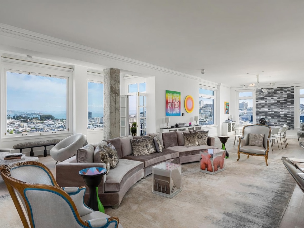 This expansive, light-filled living room features a large, plush velvet sectional sofa and a mix of modern and classic furniture pieces, including ornate armchairs. The space is defined by its panoramic city and bay views framed by large windows, a raw concrete structural column, and a neutral color palette accented by vibrant contemporary art. The open-concept layout flows seamlessly into a dining area, creating an airy and sophisticated atmosphere suitable for high-end urban living.