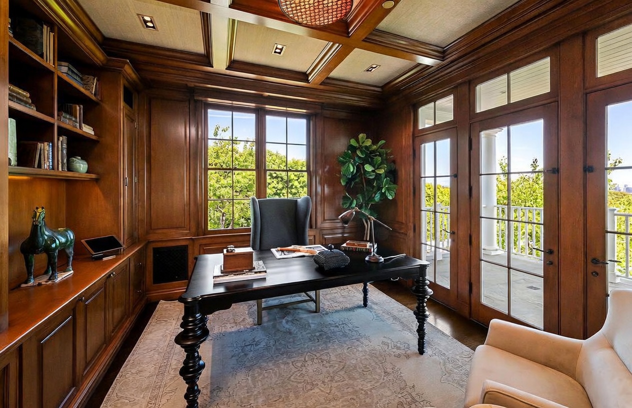 This sophisticated home office features rich wood-paneled walls, a coffered ceiling, and built-in shelving units that provide ample storage. A dark, traditional desk sits centrally on a patterned area rug, complemented by a high-backed chair and a secondary seating area. Large windows and glass-paned doors allow for abundant natural light and provide a seamless connection to the outdoor balcony.