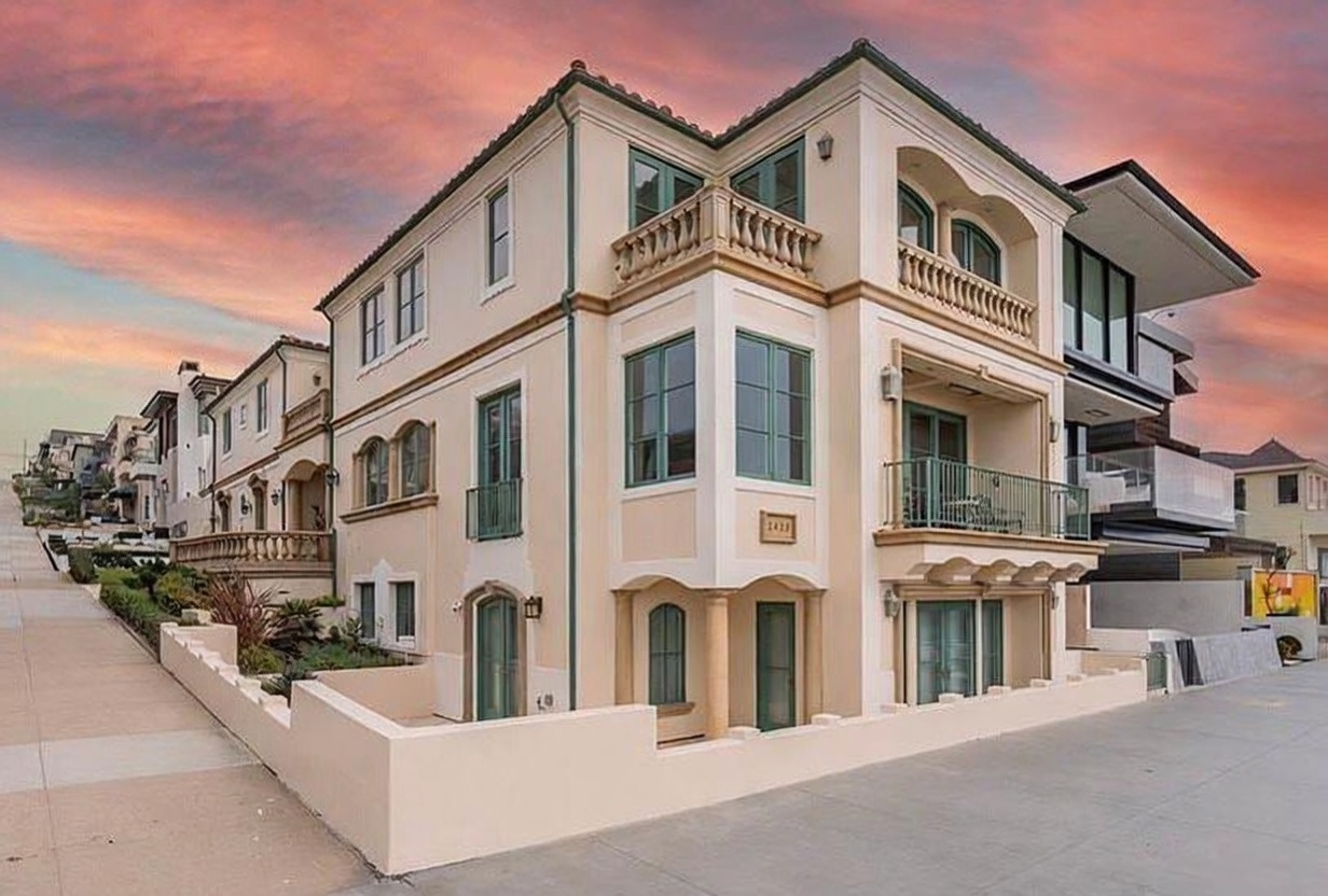This three-story Mediterranean-style residence features a light-toned stucco facade with elegant architectural details, including arched windows, decorative balustrades, and multiple balconies. The home is situated on a corner lot, showcasing a clean, sophisticated exterior design with a prominent street-level entrance and a sidewalk running along the side. The warm, sunset-hued sky provides a dramatic backdrop, highlighting the building's structured geometry and classic aesthetic.