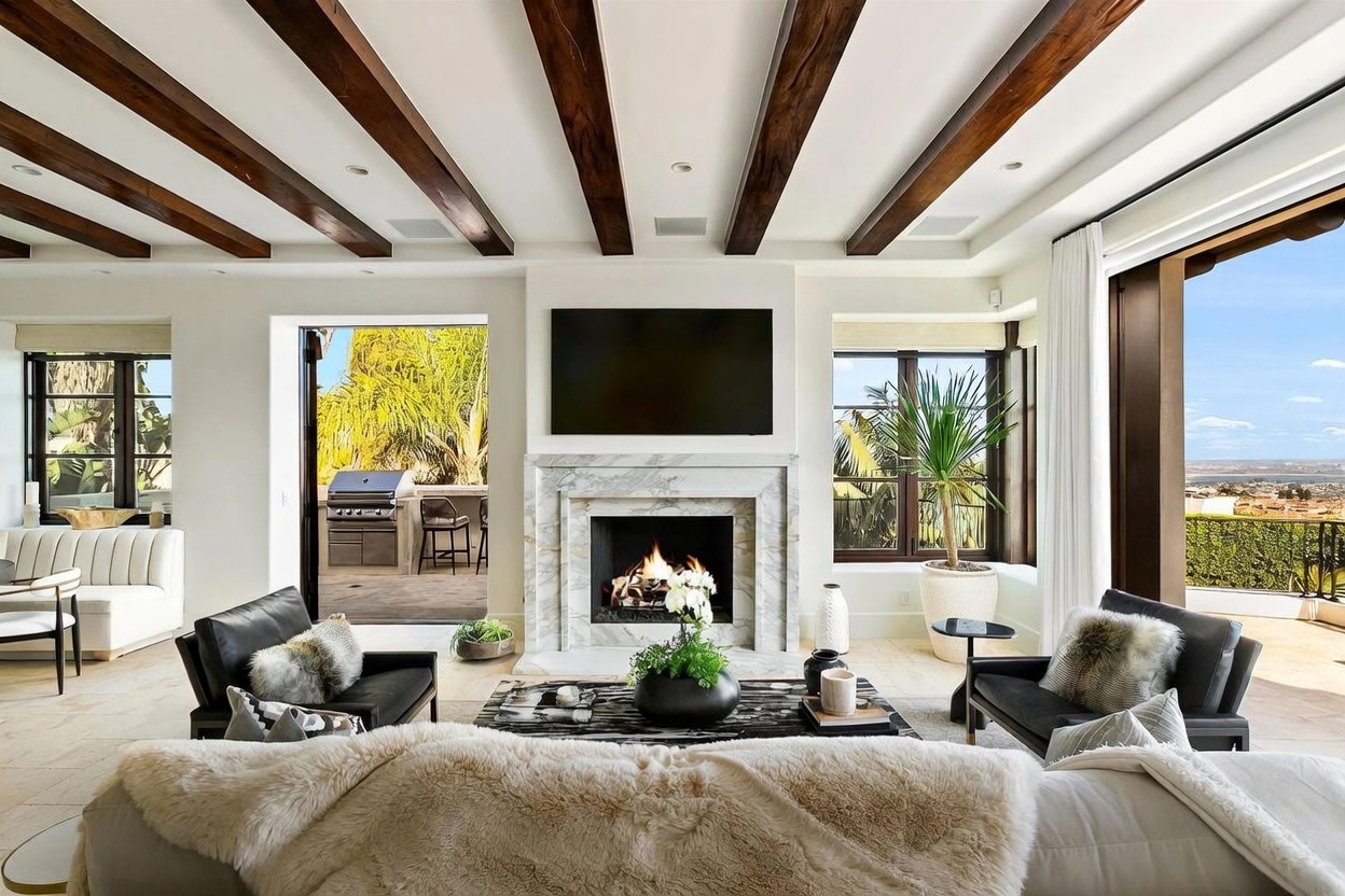 This sophisticated living room features a prominent stone-clad fireplace surround and exposed dark wood ceiling beams that add architectural character. The space is furnished with two black leather armchairs and a plush sofa, centered around a patterned coffee table, while large glass doors open to an outdoor patio area. The room is bathed in natural light, offering a seamless transition between the elegant interior and the scenic outdoor views.