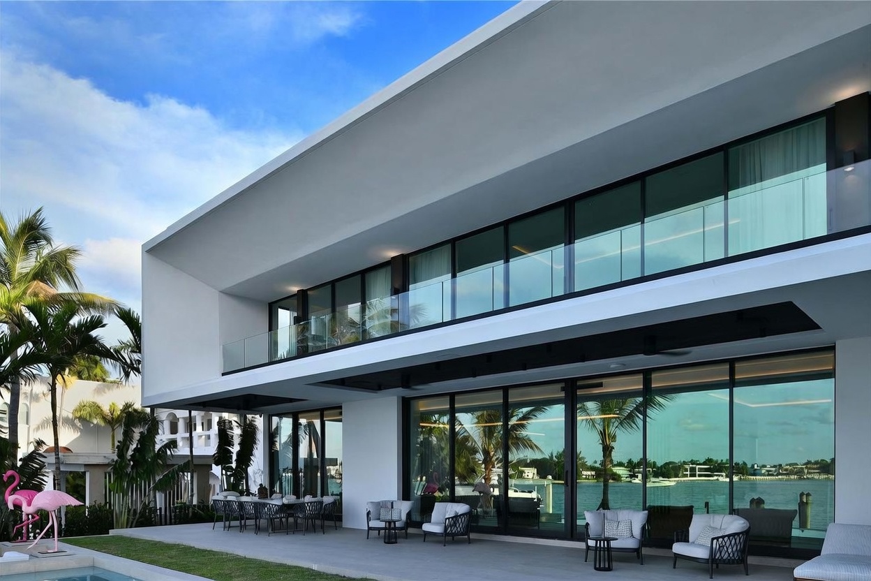 This modern, two-story luxury residence features a sleek, minimalist design with expansive floor-to-ceiling glass walls that seamlessly connect the indoor living spaces to a spacious covered patio. The outdoor area is furnished with contemporary seating and dining sets, overlooking a serene waterfront view. The architectural style emphasizes clean horizontal lines, a bright white facade, and a sophisticated, open-air aesthetic perfect for waterfront entertaining.