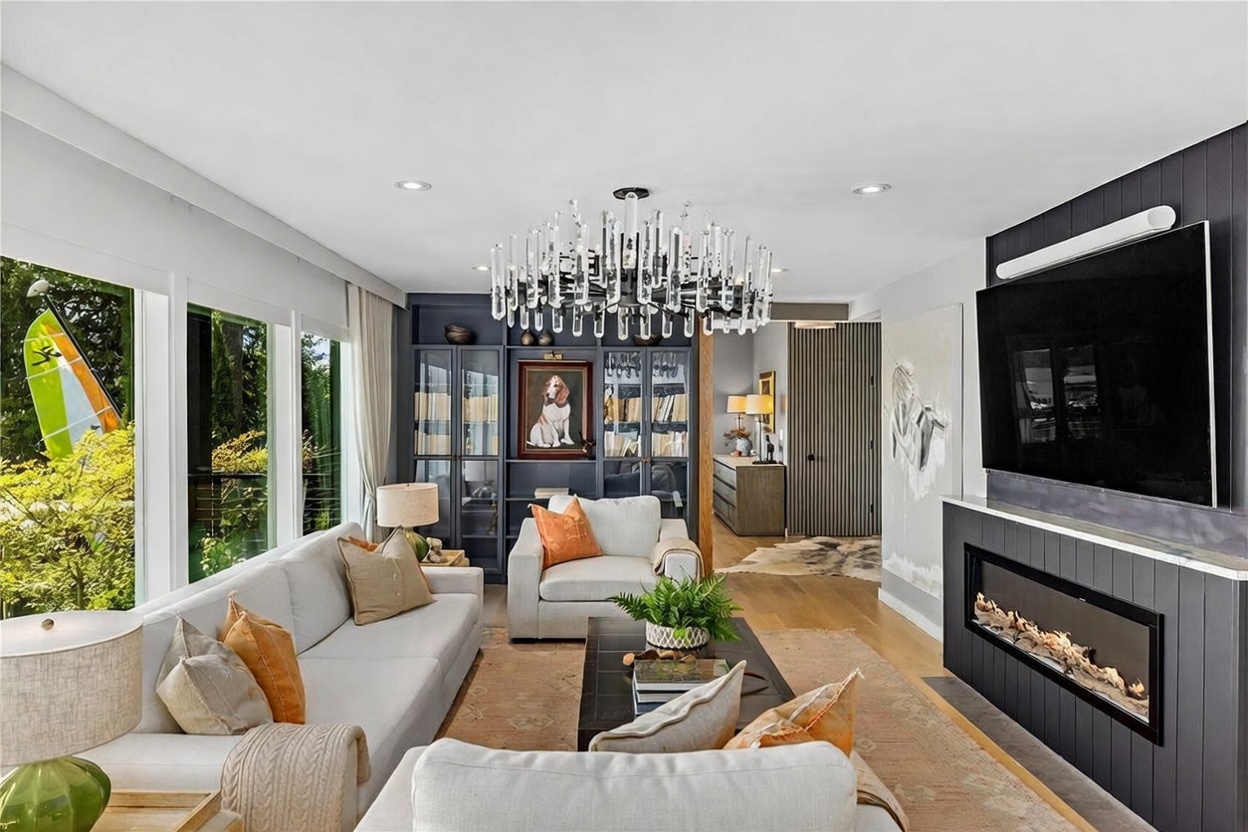 This sophisticated living room features a light-toned sofa and armchair arranged around a dark coffee table on a patterned area rug. A striking crystal chandelier serves as the focal point, while a dark-paneled wall with built-in shelving and a modern linear fireplace adds dramatic contrast. Large windows along the left wall provide ample natural light and views of the outdoors, creating an inviting and elegant atmosphere.