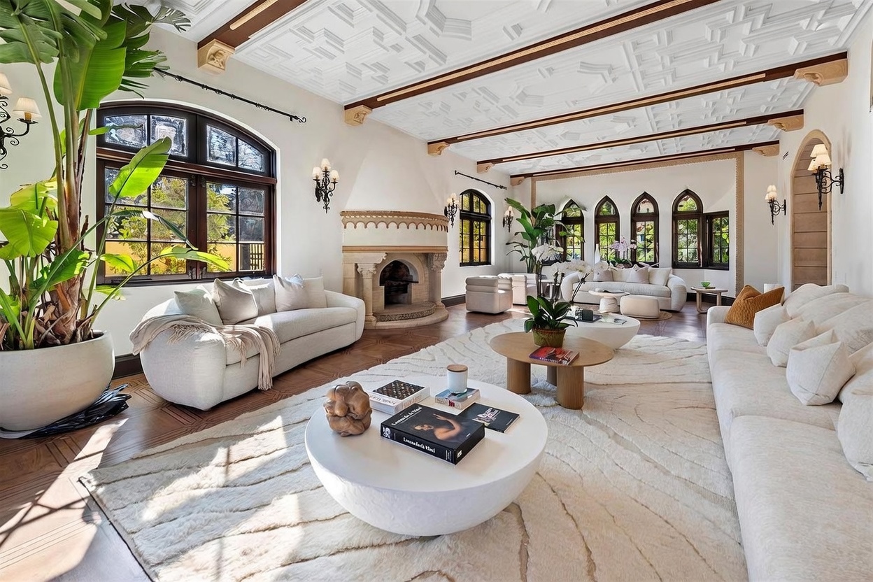 This expansive living room features a sophisticated, neutral aesthetic with a coffered ceiling and dark wood beams. A prominent stone fireplace serves as a central focal point, complemented by multiple plush sofas and a large, textured area rug. The space is bathed in natural light from several arched windows, creating an airy and elegant atmosphere suitable for formal entertaining.