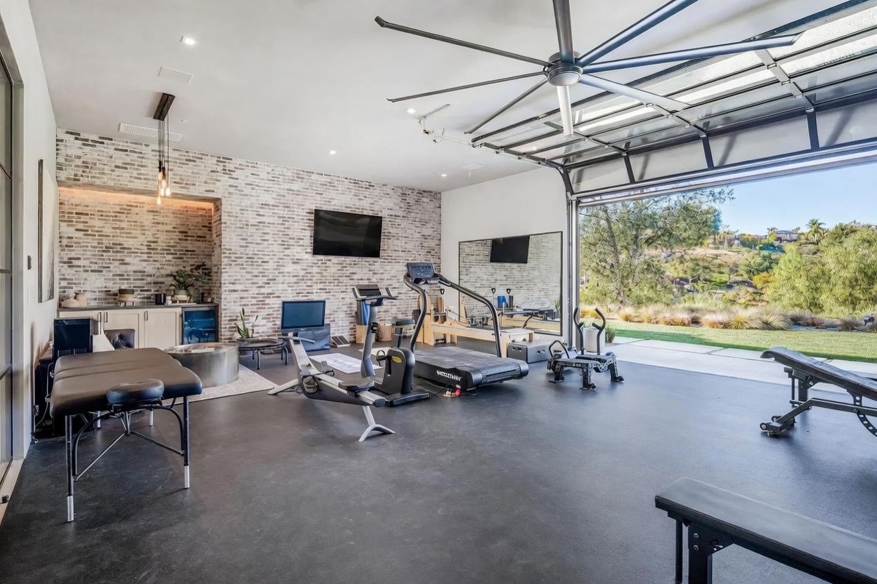 This spacious home gym features a sleek, industrial aesthetic with a dark rubberized floor and a prominent exposed brick wall. The room is equipped with professional-grade fitness equipment, including a treadmill, rowing machine, and a massage table, while a large, retractable glass garage door opens to reveal a scenic outdoor landscape. A large ceiling fan and a mirrored wall enhance the airy, functional feel of this high-end workout space.
