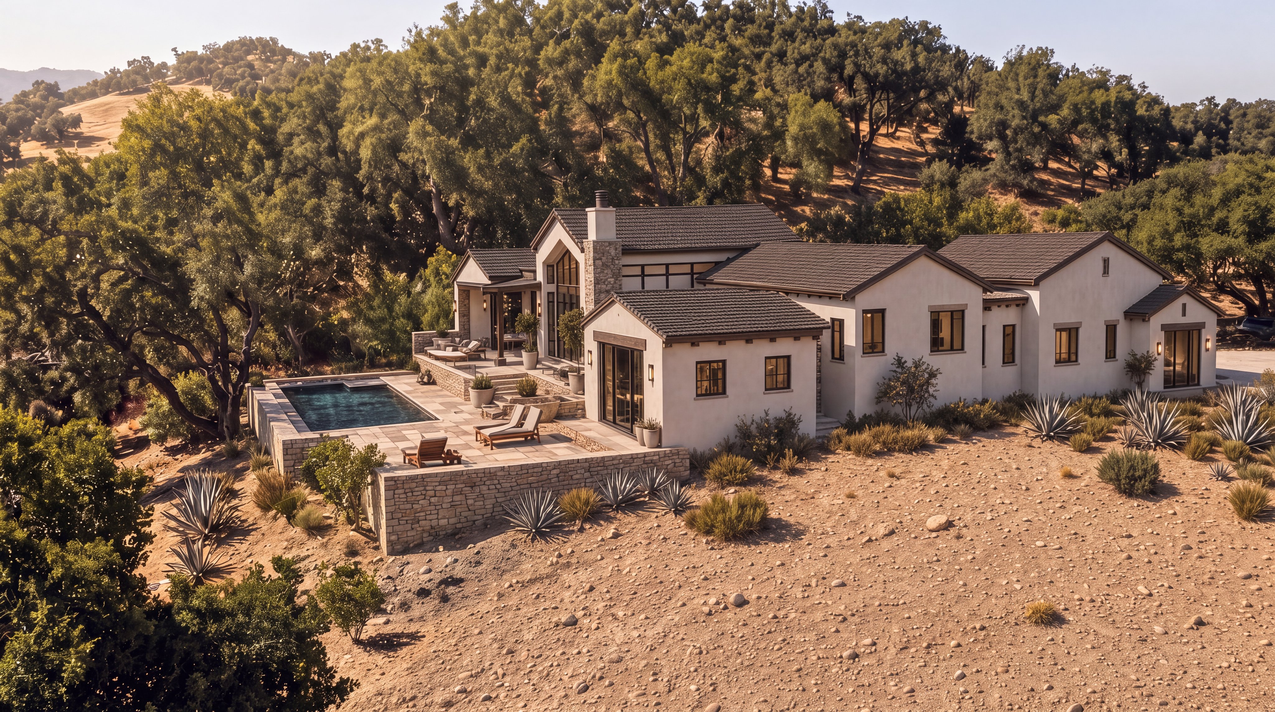 This stunning Mediterranean-style estate is captured from an elevated perspective, showcasing its harmonious integration with the surrounding hilly landscape. The property features a spacious stone patio, a sleek rectangular swimming pool, and pristine white stucco architecture complemented by dark-framed windows and a textured roof. The overall impression is one of luxury, seclusion, and sophisticated design, perfectly suited for high-end outdoor living.