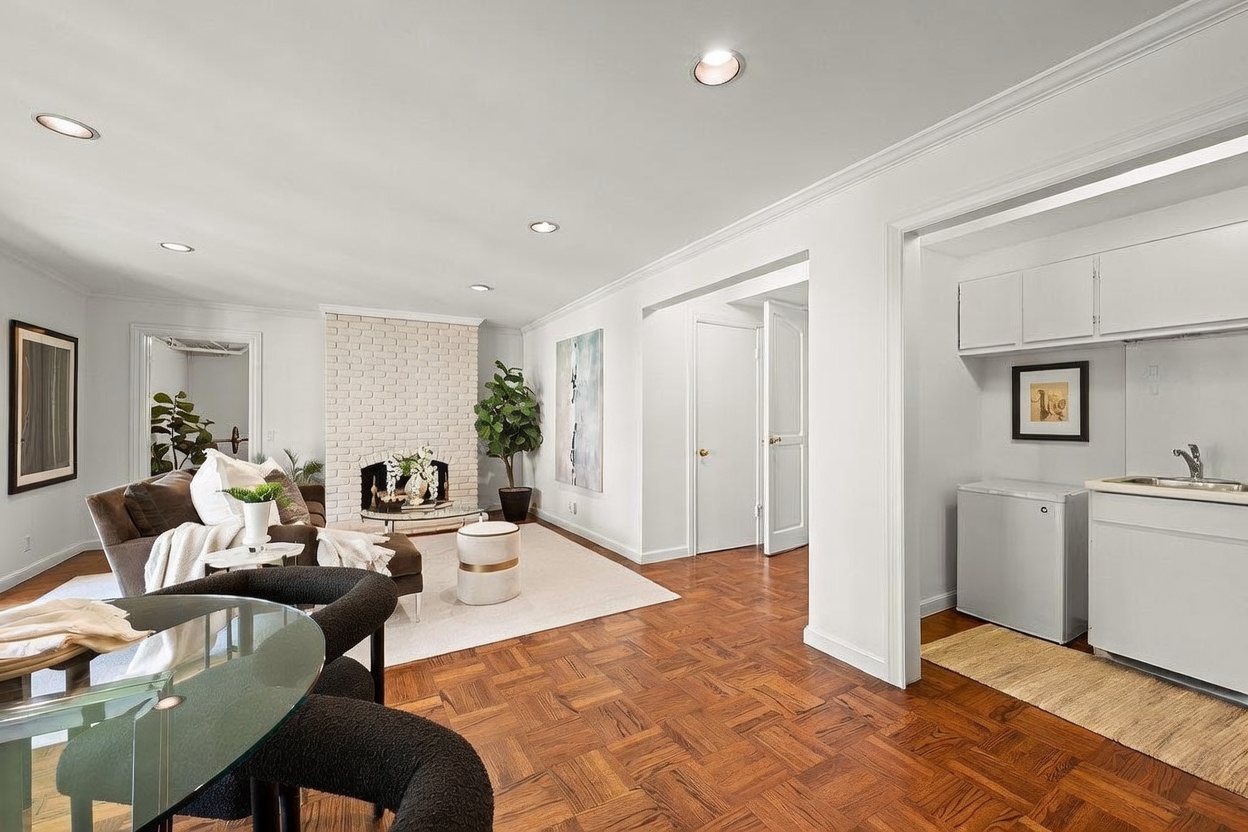This bright and open living area features a classic parquet wood floor and a prominent white brick fireplace surround. The space is furnished with a comfortable sofa, a glass-topped dining table, and a small kitchenette area tucked into an adjacent alcove. The neutral color palette and recessed lighting create a clean, inviting atmosphere suitable for both relaxation and entertaining.