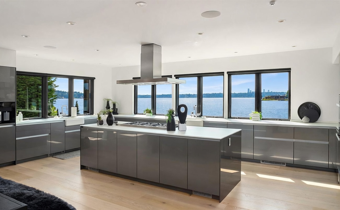 This modern, high-end kitchen features sleek, dark grey cabinetry paired with light-toned wood flooring and a large central island. A stainless steel range hood hangs above the island, which is positioned in front of a expansive wall of windows offering a panoramic view of the water. The space is bright and airy, characterized by a minimalist aesthetic and clean lines that emphasize the connection to the outdoor scenery.