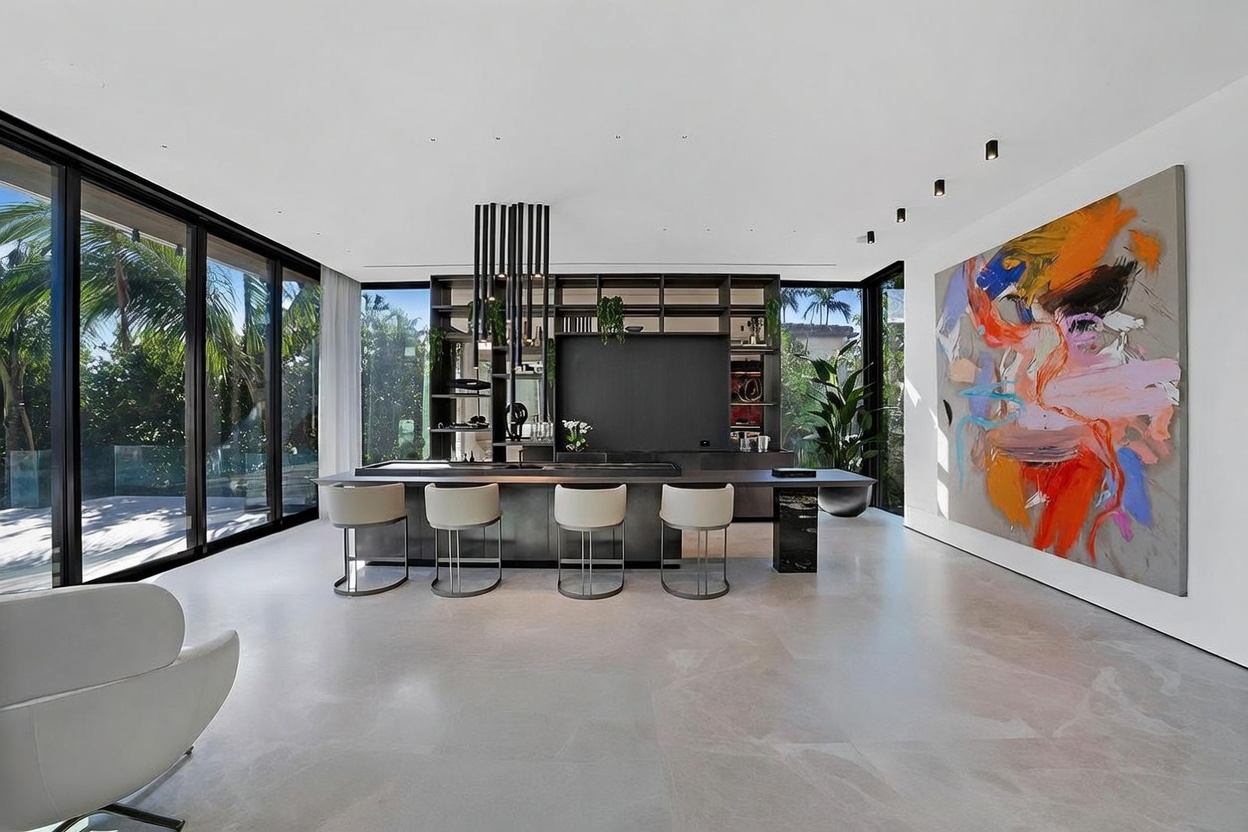 This sleek, modern kitchen features a long, dark-toned island with integrated seating for four, positioned beneath a striking arrangement of vertical pendant lights. The space is defined by its minimalist aesthetic, including dark cabinetry with open shelving, a built-in television, and expansive floor-to-ceiling windows that provide abundant natural light and views of the lush greenery outside. A large, vibrant abstract painting adds a bold splash of color to the otherwise neutral, contemporary interior.