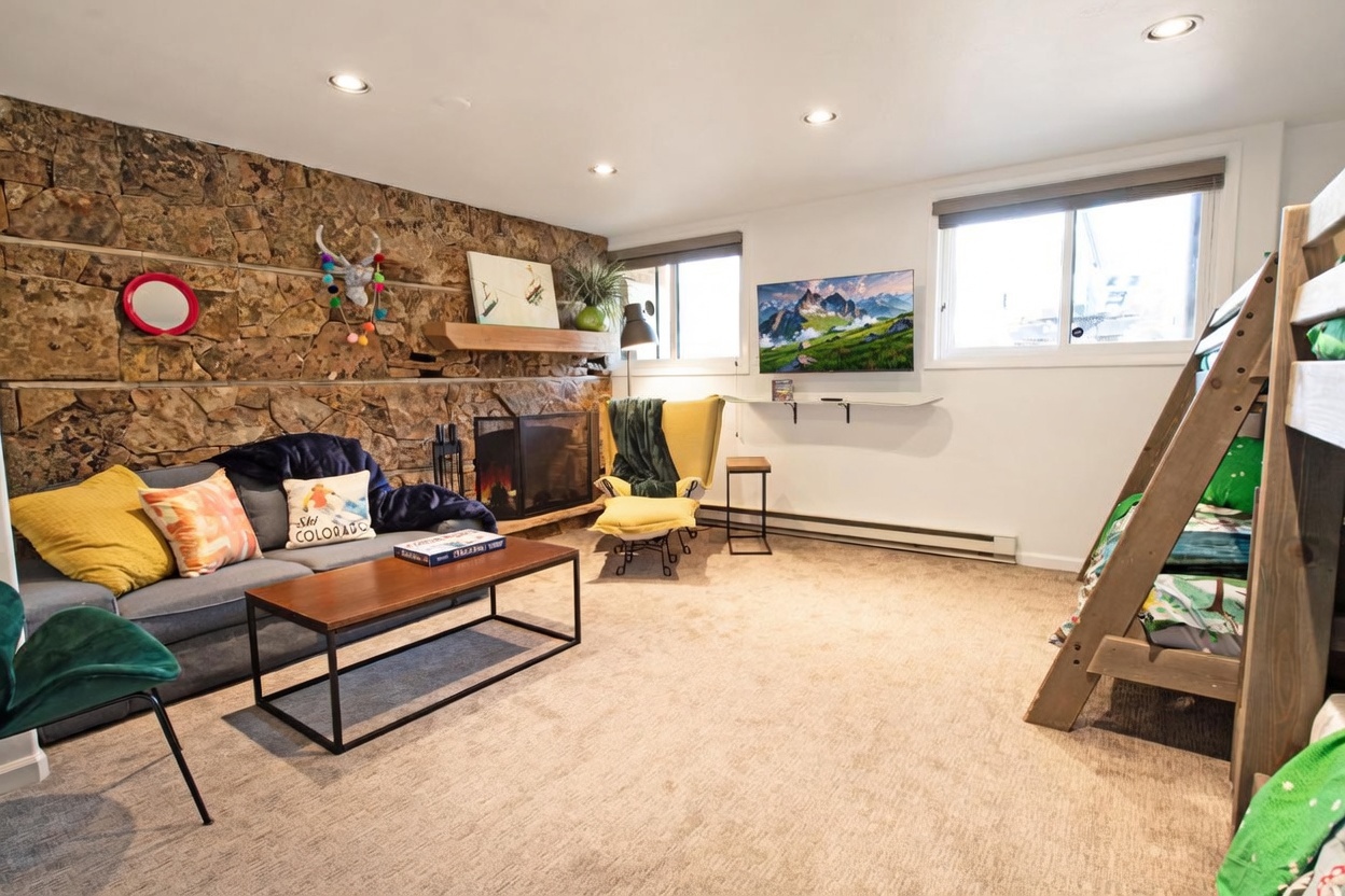 This cozy living room features a prominent stone-clad wall with a built-in fireplace and a wooden mantel, creating a rustic mountain aesthetic. The space is furnished with a grey sofa, a yellow accent chair with an ottoman, and a wooden coffee table, while a bunk bed is positioned to the right. Large windows allow natural light to brighten the room, which is finished with neutral carpeting and recessed lighting.