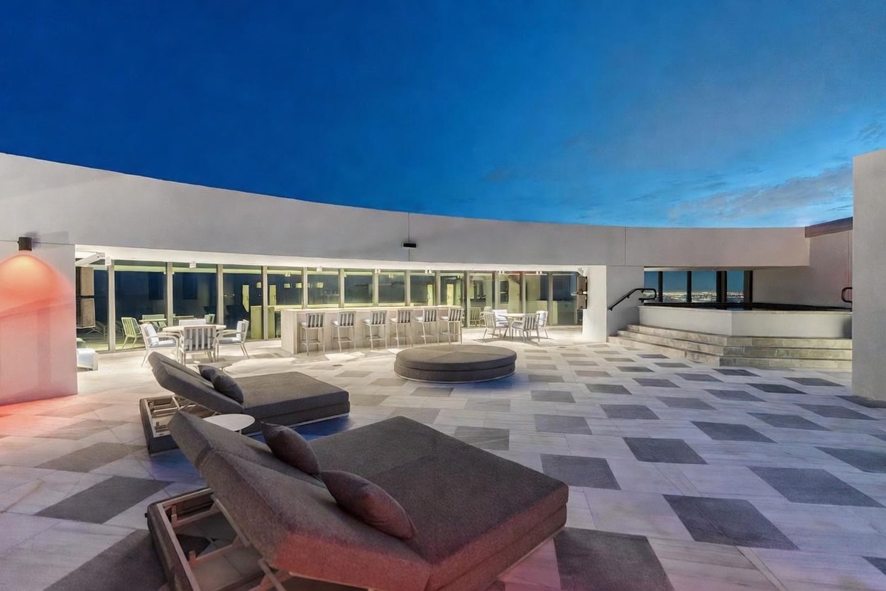This expansive rooftop terrace features a modern, open-air design with a checkered tile floor and panoramic views. The space is furnished with multiple lounge chairs, a circular daybed, and a bar area with high stools, all set against a backdrop of glass walls and a twilight sky. A raised platform with steps leads to a spa area, creating a sophisticated and inviting atmosphere for outdoor entertaining.