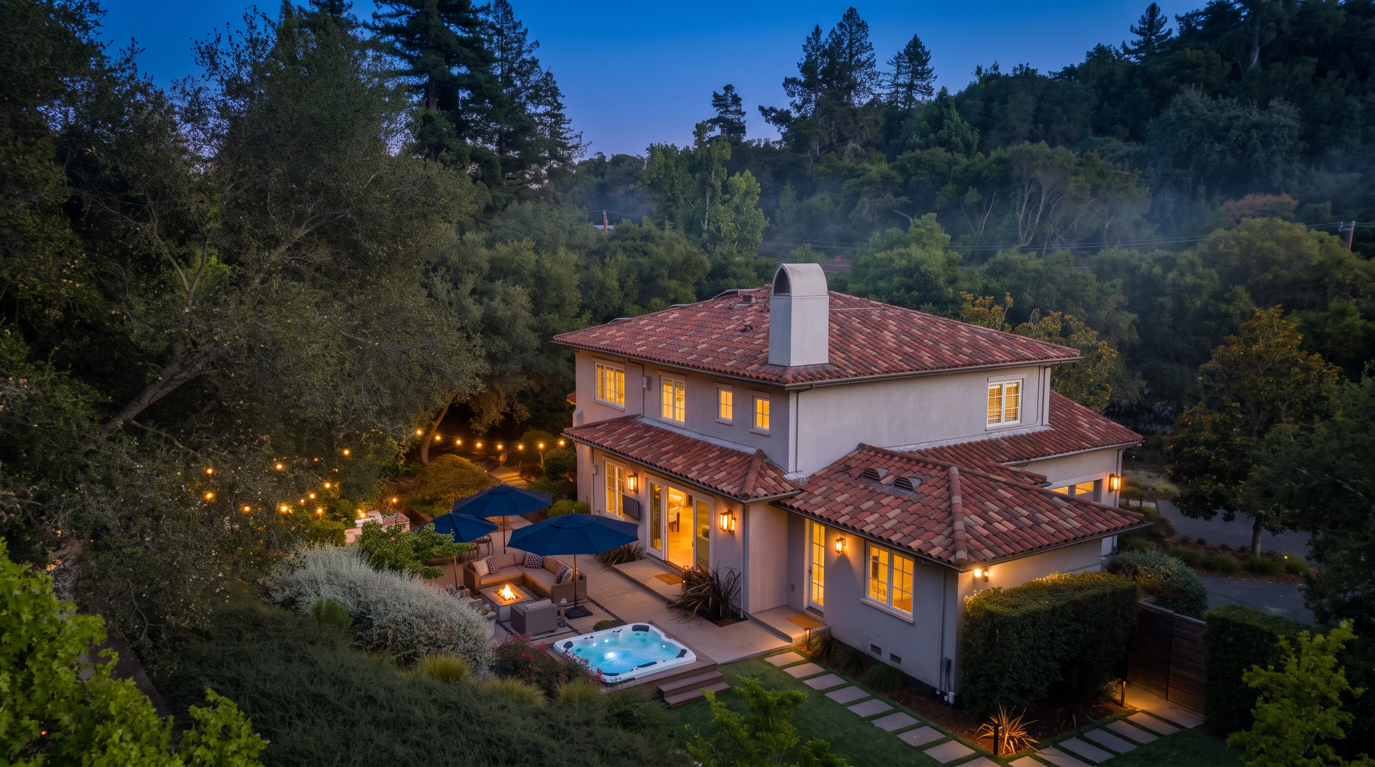 This aerial perspective captures a two-story Mediterranean-style home with a distinctive terracotta tile roof and light-colored stucco exterior. The backyard features a well-appointed patio area with outdoor seating, blue umbrellas, and a private spa, all nestled against a lush, wooded backdrop. A manicured lawn with a stone paver path completes the serene outdoor living space, creating an inviting atmosphere for relaxation and entertaining.