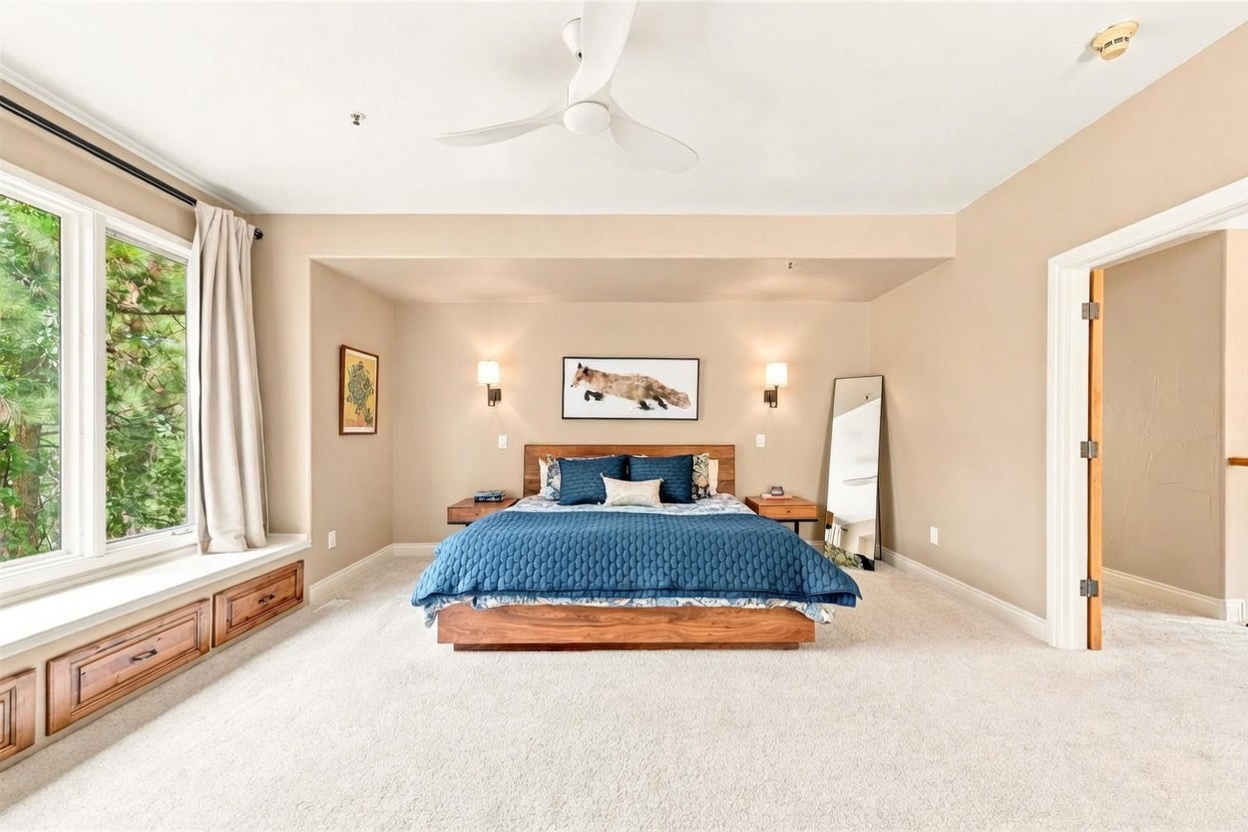 This spacious primary bedroom features a central bed with a dark blue quilted coverlet and a wood frame, flanked by matching wall-mounted nightstands and sconces. A large window with a built-in bench and storage drawers provides a view of the surrounding trees, while a full-length mirror stands in the corner. The room is finished with neutral-toned walls and plush carpeting, creating a calm and inviting atmosphere.