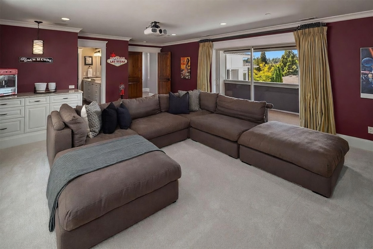 This spacious media room features deep burgundy walls and a large, plush sectional sofa arranged for comfortable viewing. A projector is mounted on the ceiling, and a built-in cabinet with a popcorn machine adds a fun, theater-like atmosphere to the space. Large sliding glass doors provide access to a balcony, allowing natural light to brighten the room while offering a view of the surrounding trees.