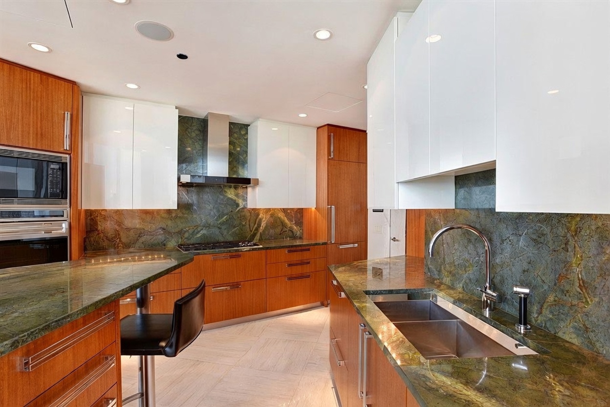 This modern kitchen features a striking contrast between warm-toned wood cabinetry and sleek white upper cabinets. The space is anchored by dramatic green stone countertops and a matching full-height backsplash, complemented by stainless steel appliances and a double-basin sink. The perspective captures the functional layout, including a breakfast bar with a black stool and recessed ceiling lighting, creating a sophisticated and high-end culinary environment.