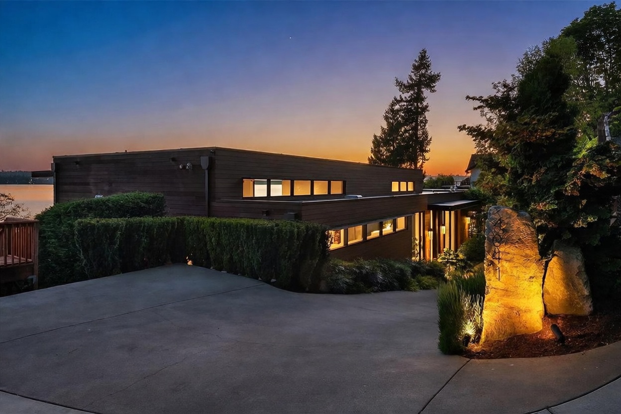 This contemporary home is captured at dusk, showcasing its sleek, horizontal wood-clad architecture and warm, inviting exterior lighting. A concrete driveway leads toward the entrance, which is marked by a large, illuminated boulder featuring the house number. The scene is framed by lush landscaping and a sunset sky, creating a sophisticated and tranquil atmosphere.