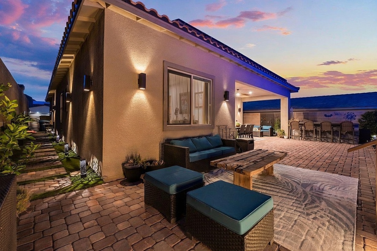 This inviting outdoor living space features a covered patio area with a comfortable sofa, two ottomans, and a rustic wooden coffee table set atop a large area rug. The space is paved with stone pavers and extends into a side yard with a landscaped walkway, all illuminated by warm wall-mounted lighting. The scene is captured at dusk, highlighting the cozy atmosphere and the transition between the indoor and outdoor living areas.
