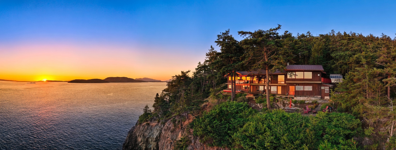 This stunning aerial perspective captures a contemporary wood-sided home perched dramatically on a rugged cliffside overlooking a vast, tranquil bay at sunset. The house features expansive glass windows that reflect the golden hour light, while the surrounding landscape is densely forested with evergreen trees. A small patio area with outdoor seating sits near the edge of the cliff, emphasizing the property's unique and secluded coastal location.