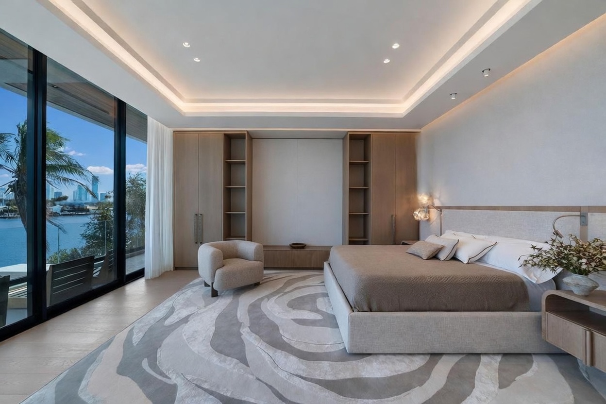 This sophisticated primary bedroom features a neutral color palette and a large area rug with a swirling, organic pattern. The room is anchored by a plush bed and a curved accent chair, while a wall of custom light wood cabinetry provides integrated storage and open shelving. Floor-to-ceiling windows offer expansive views of the water and city skyline, bathing the space in natural light.