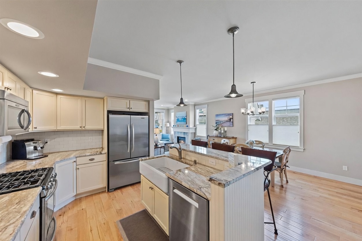 This bright, open-concept kitchen features light-toned cabinetry, granite countertops, and stainless steel appliances, including a farmhouse-style sink integrated into the central island. The space flows seamlessly into a dining area and a living room with a fireplace, all tied together by warm-toned wood flooring. The perspective is from the kitchen, looking past the island toward the dining and living areas, creating an inviting and spacious atmosphere.