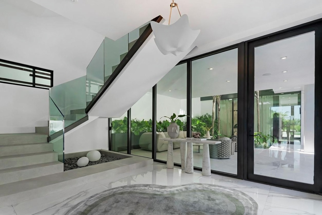 This modern entryway features a striking staircase with glass railings and a sculptural pendant light suspended above. A stone console table sits against a glass wall that looks into a lush, plant-filled interior courtyard, while a smooth, curved rug anchors the polished white floor. The space is defined by its clean lines, minimalist aesthetic, and seamless integration of indoor and outdoor elements.