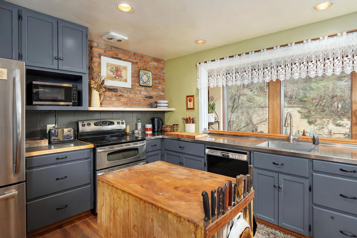 This rustic-inspired kitchen features blue-painted cabinetry paired with a prominent exposed brick wall and a large, weathered wood butcher block island. A stainless steel stove and dishwasher are integrated into the workspace, which is illuminated by recessed lighting and natural light streaming through a large window adorned with a lace valance. The space offers a warm, lived-in aesthetic with a functional layout that includes a deep stainless steel sink and open shelving.