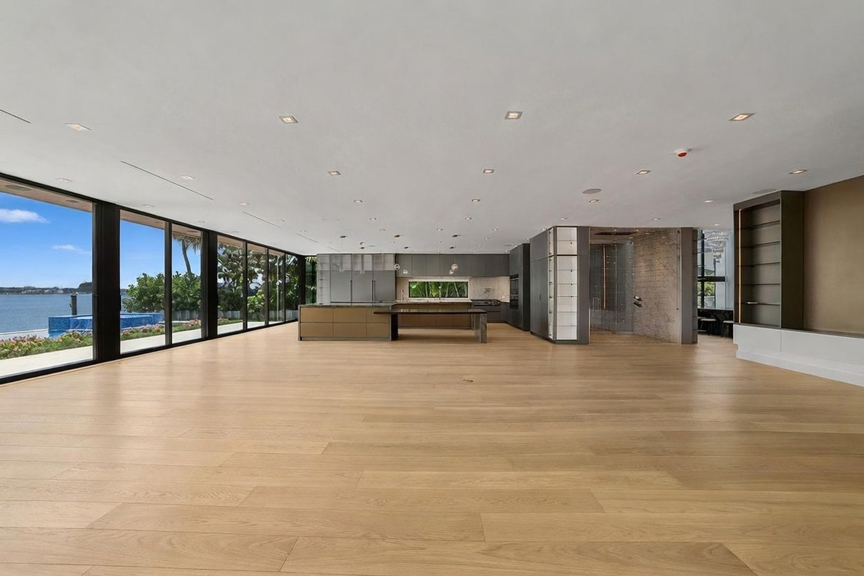 This expansive, open-concept living area features a sleek, modern kitchen with a large central island and dark cabinetry. The space is defined by light wood flooring and a wall of floor-to-ceiling glass that provides an unobstructed view of the outdoor pool and waterfront. The minimalist design emphasizes clean lines and a seamless transition between the indoor living space and the exterior environment.