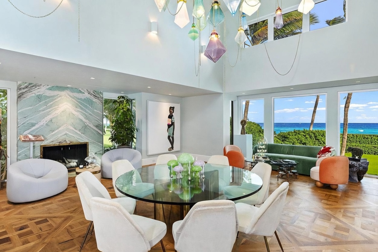 This expansive, high-ceilinged dining area features a large circular glass table surrounded by modern upholstered chairs, positioned beneath a dramatic, multi-colored geometric chandelier. To the left, a striking green-veined stone fireplace surround serves as a focal point, while the right side of the room opens up to panoramic ocean views through large windows. The space is finished with intricate parquet wood flooring and decorated with contemporary art, creating a sophisticated and airy atmosphere.