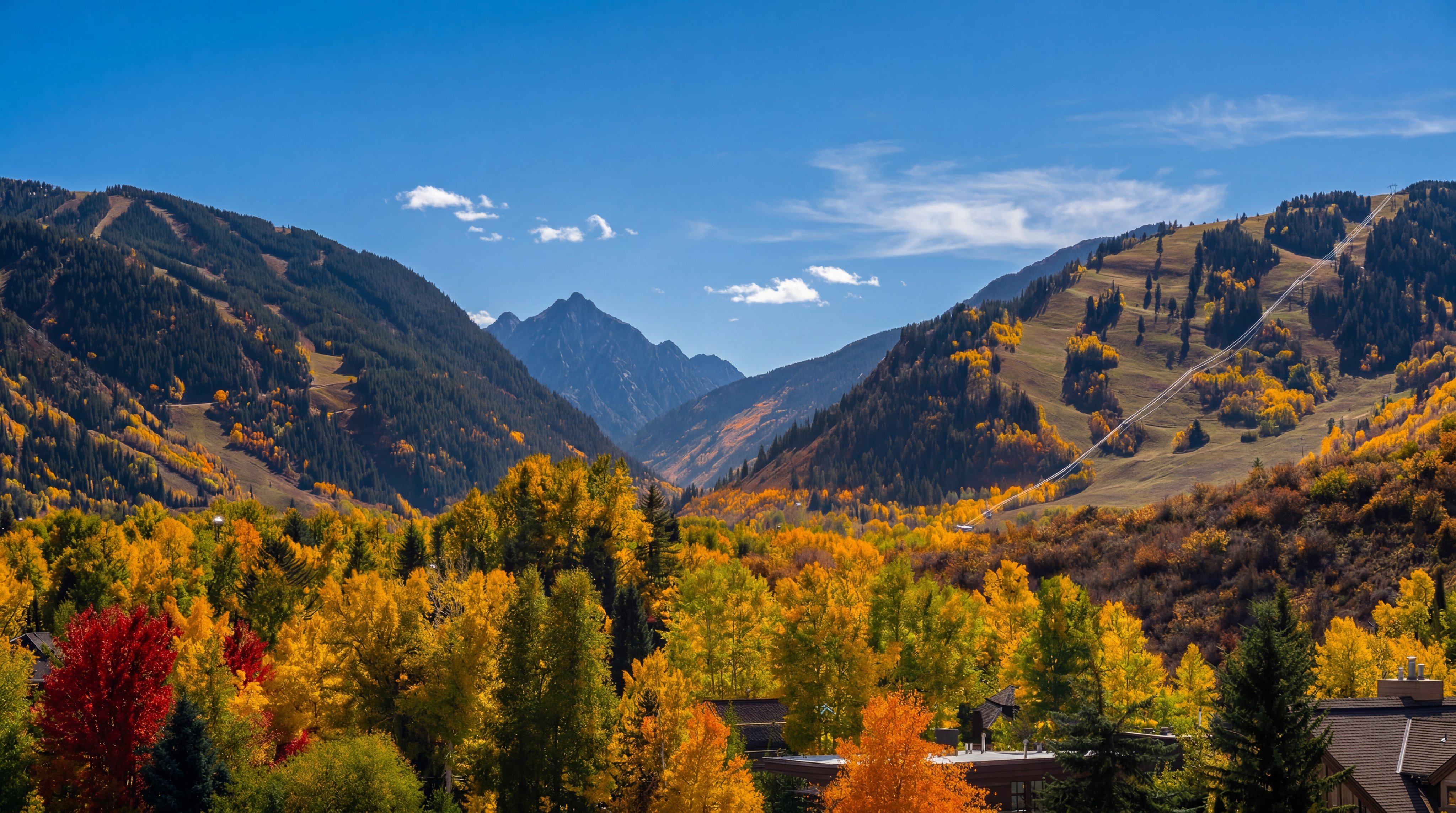 This elevated perspective captures a breathtaking mountain landscape during the peak of autumn, showcasing a vibrant tapestry of golden and orange foliage. The scene features dramatic, rugged mountain peaks in the distance, framed by a clear blue sky with scattered white clouds. In the foreground, the tops of trees and a glimpse of a residential roof structure suggest a scenic, high-altitude setting ideal for nature enthusiasts.
