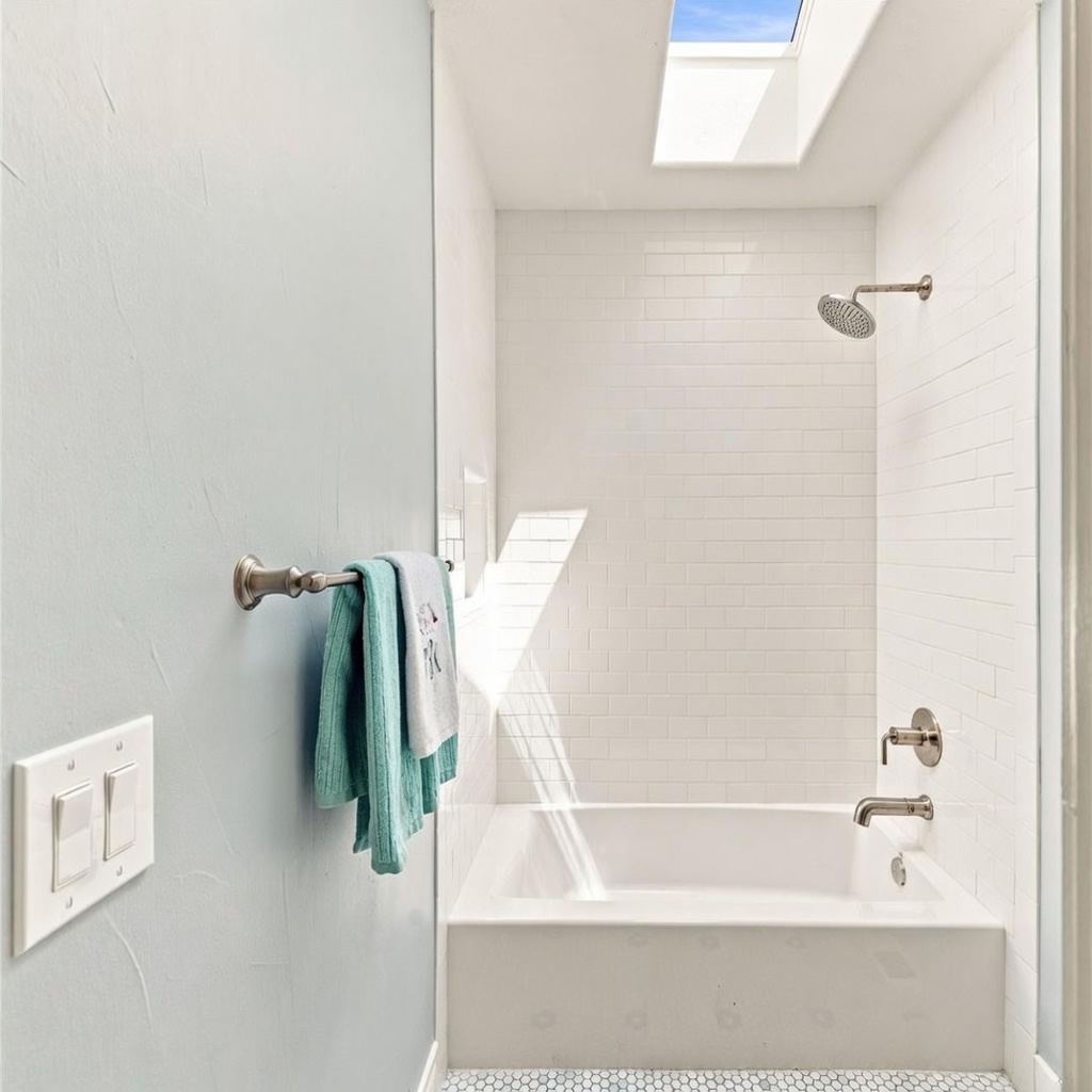 This bright bathroom features a clean, minimalist aesthetic with a white subway-tiled shower surround and a built-in bathtub. A skylight above the shower area floods the space with natural light, highlighting the light blue wall and the modern brushed-nickel fixtures. The perspective is a straight-on shot looking into the shower alcove, emphasizing the airy and well-lit design.