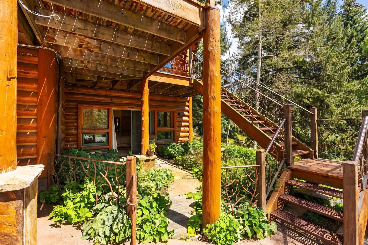 This image captures a rustic log cabin exterior featuring a covered stone patio nestled beneath a wooden deck. A distinctive metal staircase with intricate, branch-like railings provides access to the upper level, while lush green foliage and garden beds surround the base of the home. The perspective is from the ground level, highlighting the natural textures of the log walls and the organic design of the outdoor living space.