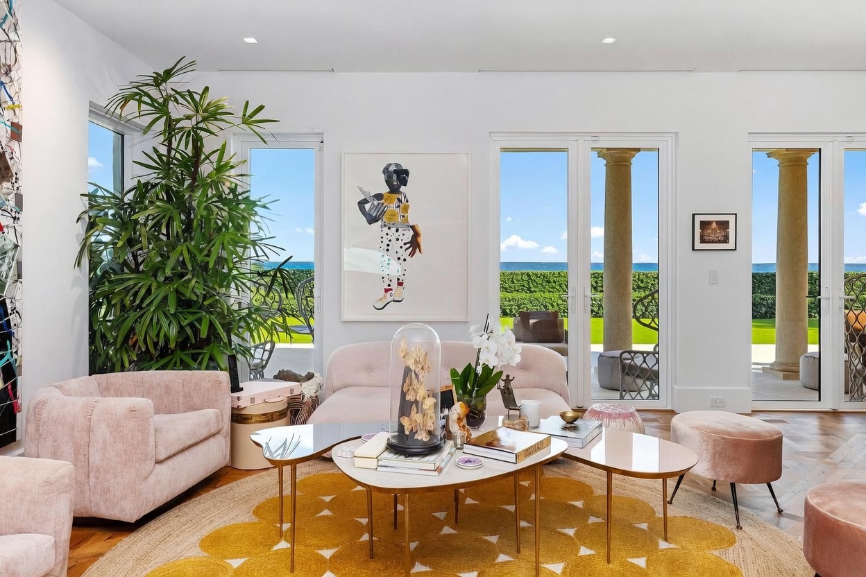 This bright and airy living room features a sophisticated, contemporary design with a soft color palette of blush pinks and warm yellows. The space is anchored by a large, circular patterned rug and a multi-tiered coffee table, complemented by plush seating and a tall indoor plant. Large glass doors provide an expansive view of the ocean and a manicured hedge, creating a seamless connection between the elegant interior and the coastal landscape.
