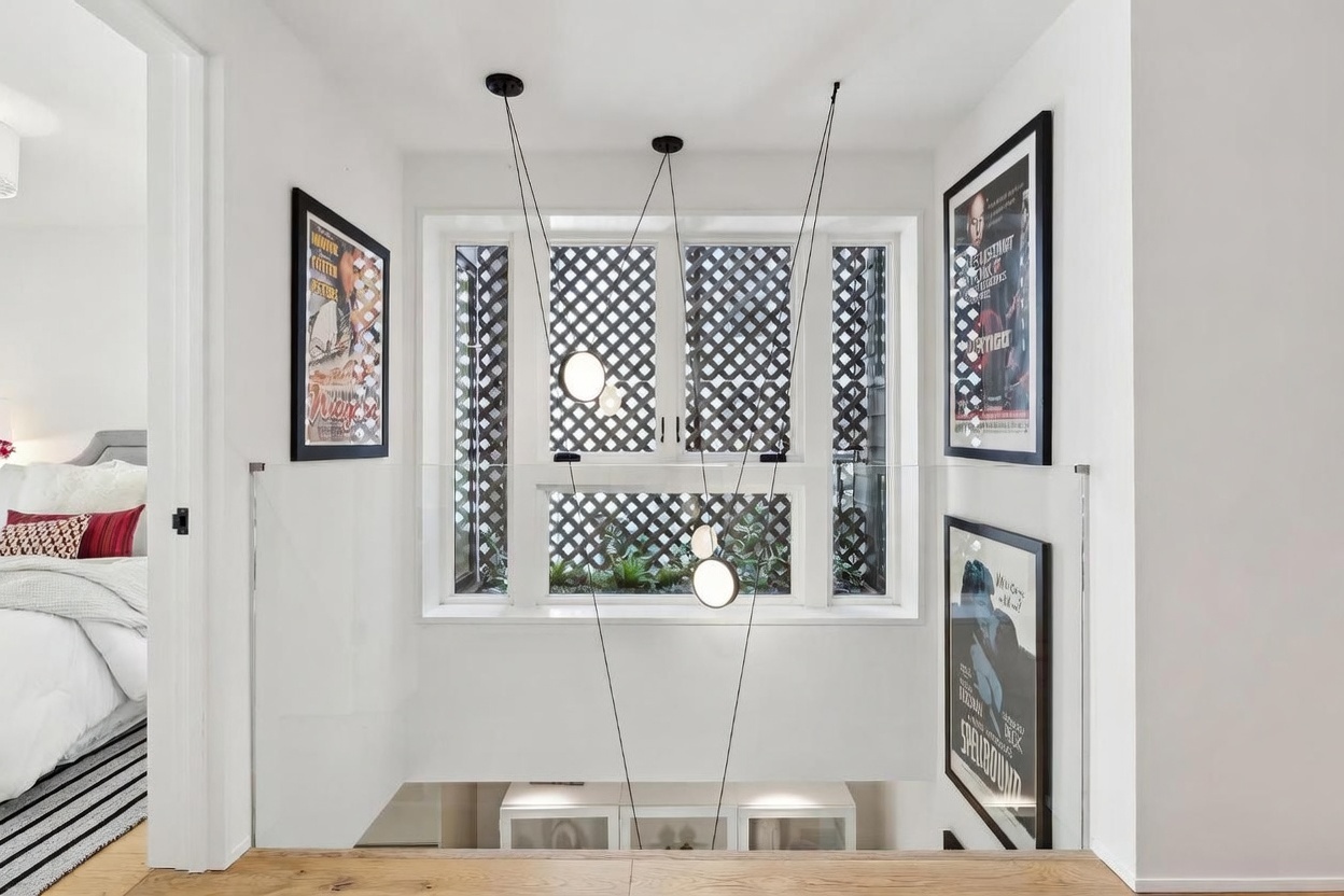 This bright landing area features a modern, multi-pendant light fixture suspended in front of a large window with a decorative lattice screen. To the left, a glimpse of a bedroom with a patterned accent pillow is visible, while the walls are adorned with framed vintage movie posters. The space is defined by clean white walls, light wood flooring, and a glass railing that overlooks the level below.