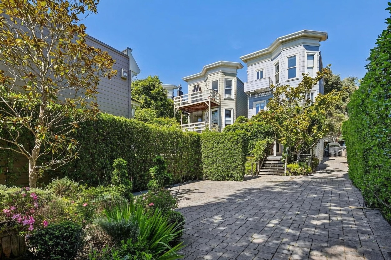 This rear view of a multi-story residential property showcases a paved driveway leading toward a set of stairs and a lush, manicured garden. The architecture features classic bay windows and a wooden balcony, framed by tall, dense hedges that provide significant privacy. The scene is captured from a low angle, emphasizing the depth of the driveway and the inviting, verdant outdoor space.