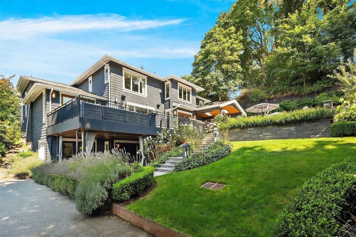 This rear view of a multi-story home showcases a dark-shingled exterior with a prominent elevated deck featuring black metal railings. A stone retaining wall borders a lush, manicured lawn that leads up to a covered patio area, creating a tiered outdoor living space. The scene is captured from a low angle on a gravel driveway, emphasizing the home's integration into the sloped, wooded landscape.