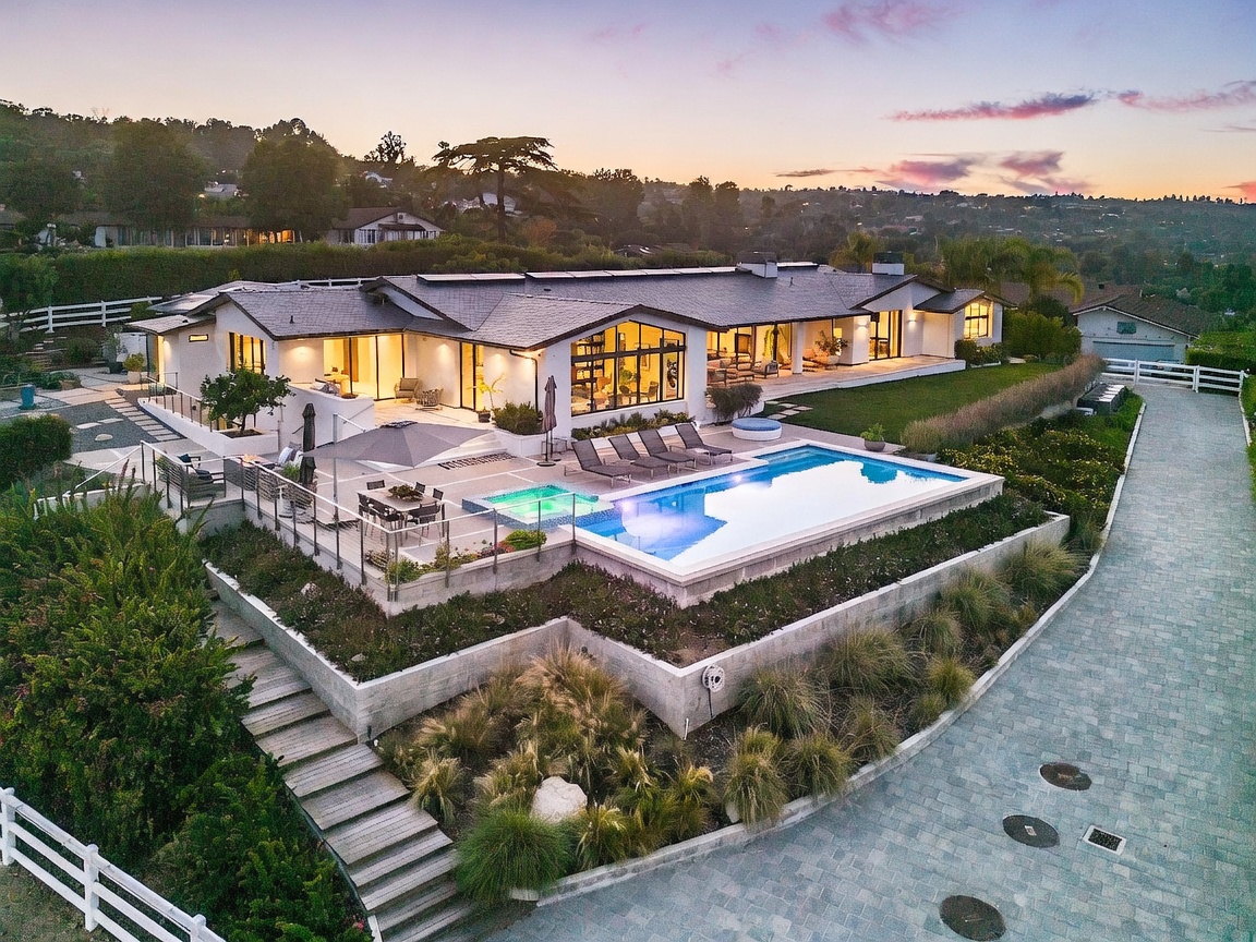 This elevated aerial perspective captures a sprawling, modern single-story residence set against a scenic hillside backdrop at dusk. The property features an expansive stone-paved patio with a rectangular swimming pool, a built-in spa, and multiple lounge areas, all overlooking a terraced landscape. The home's architecture emphasizes indoor-outdoor living with large glass panels and a clean, contemporary aesthetic.