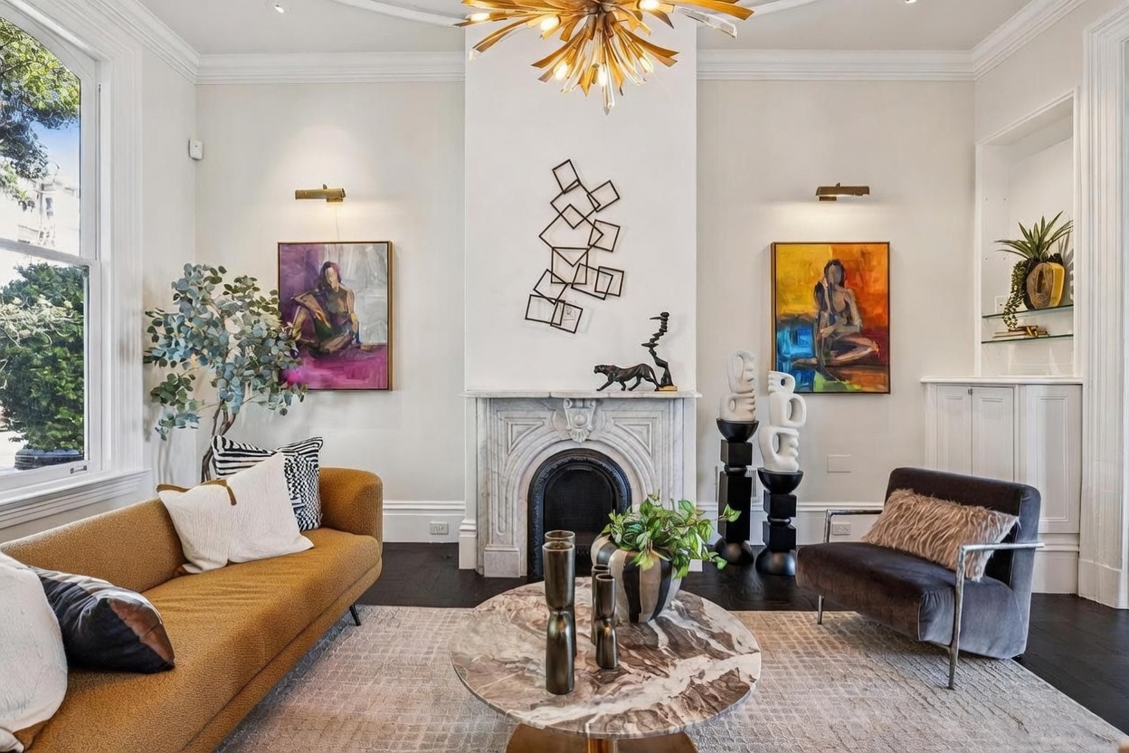 This sophisticated living room features a central fireplace with an ornate stone surround and a tall chimney breast adorned with geometric metal wall art. The space is furnished with a mustard-toned sofa, a dark velvet armchair, and a round stone-topped coffee table, all arranged on a textured area rug. Large windows on the left provide natural light, while a striking gold starburst chandelier serves as a focal point above the seating area.