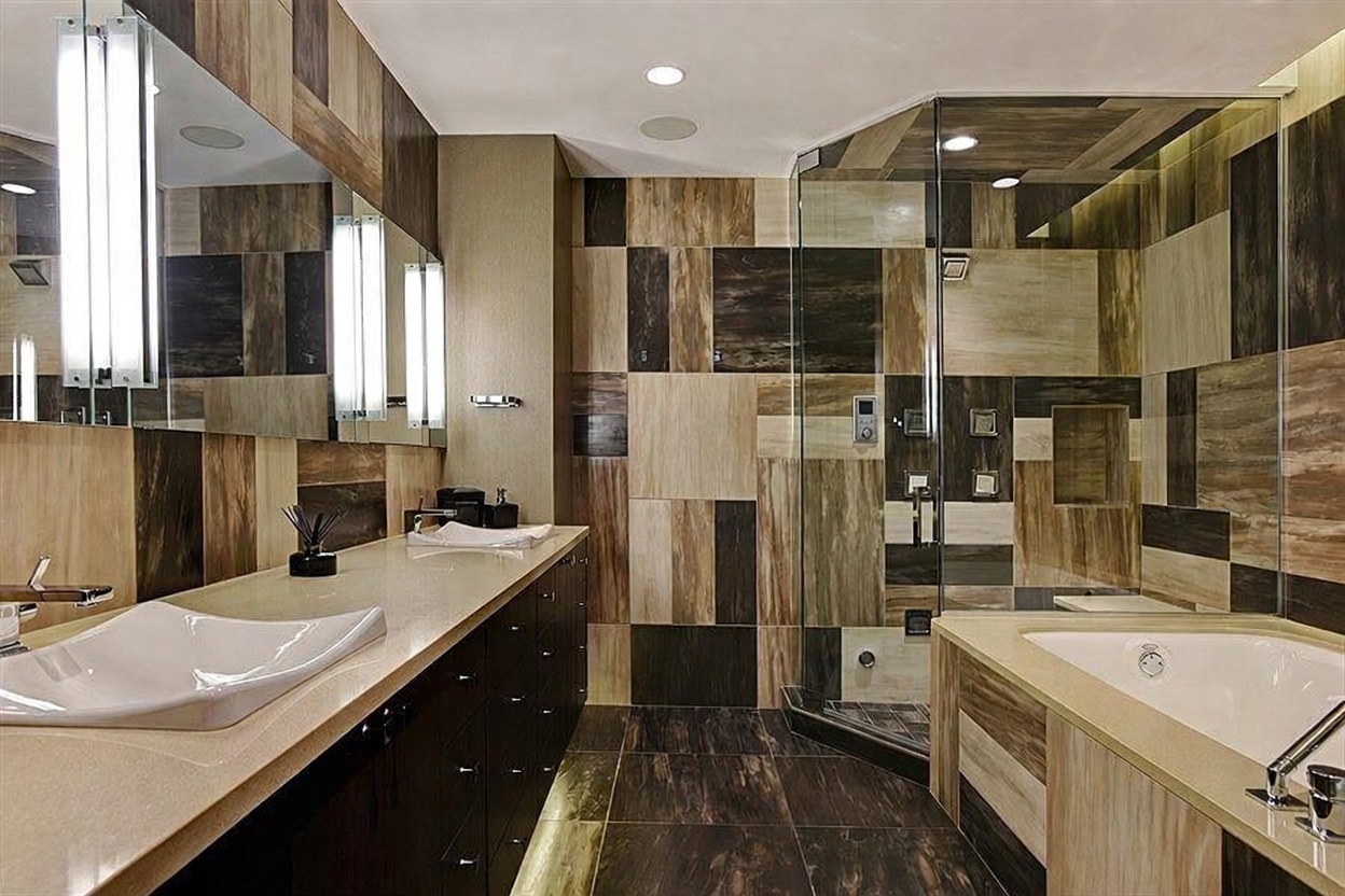 This modern primary bathroom features a dual-sink vanity with dark cabinetry and a light-toned countertop, complemented by a large mirror with integrated vertical lighting. A glass-enclosed shower stall with patterned tile walls sits adjacent to a built-in soaking tub, creating a cohesive and luxurious aesthetic. The room is finished with dark, large-format floor tiles that contrast with the multi-toned wall tiles, providing a sophisticated and contemporary atmosphere.