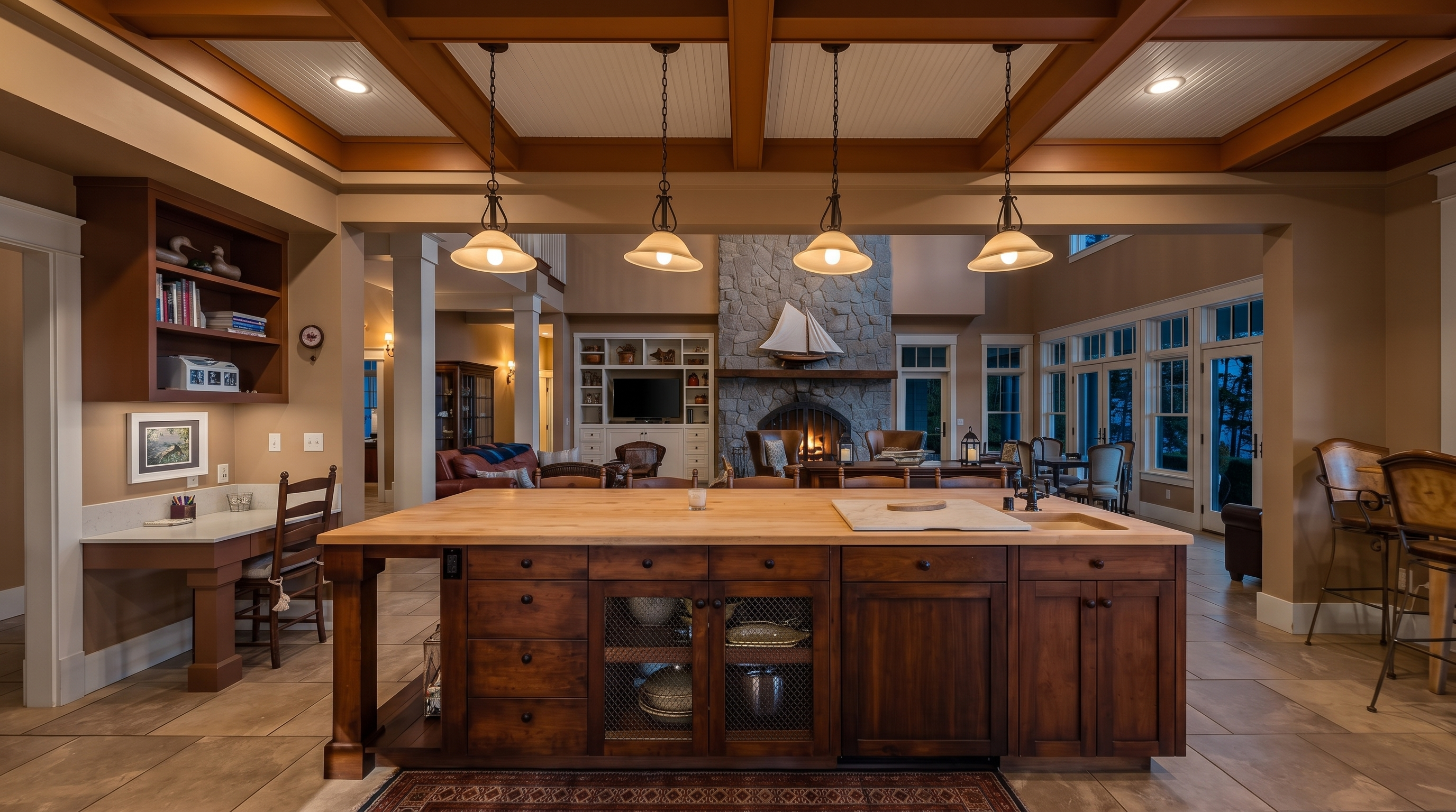 This spacious kitchen features a large central island with a light wood countertop and a built-in sink, serving as the focal point of the open-concept living area. The room is defined by a coffered ceiling with pendant lighting and a stone-clad chimney breast in the background, creating a warm and inviting atmosphere. A dedicated desk area is tucked into the side, while large windows provide ample natural light and views of the outdoors.