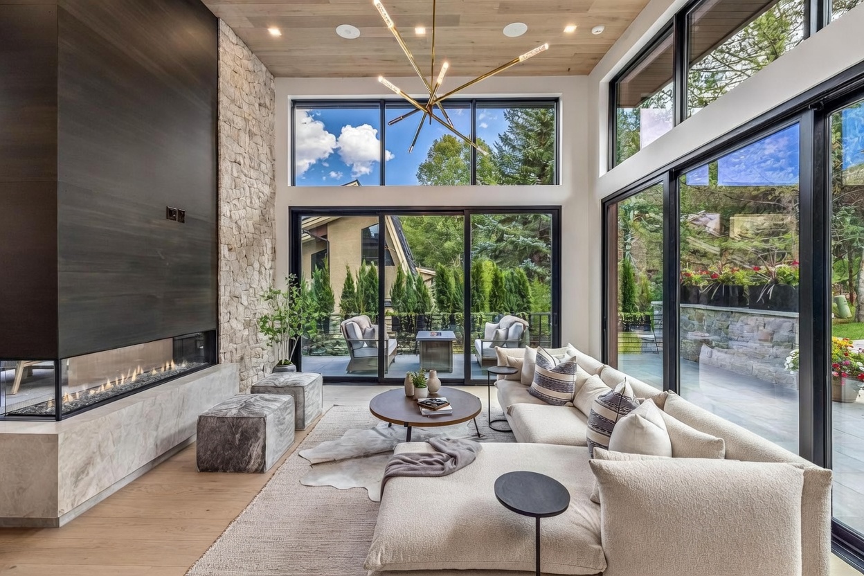 This bright and airy living room features a contemporary design with a light wood ceiling and expansive glass walls that provide a seamless connection to the outdoor patio. A prominent stone-clad chimney breast anchors the space, complemented by a long, linear fireplace set into a stone hearth. The room is furnished with a plush, neutral-toned sectional, a round coffee table, and two textured ottomans, all arranged on a large area rug to create a sophisticated and inviting atmosphere.