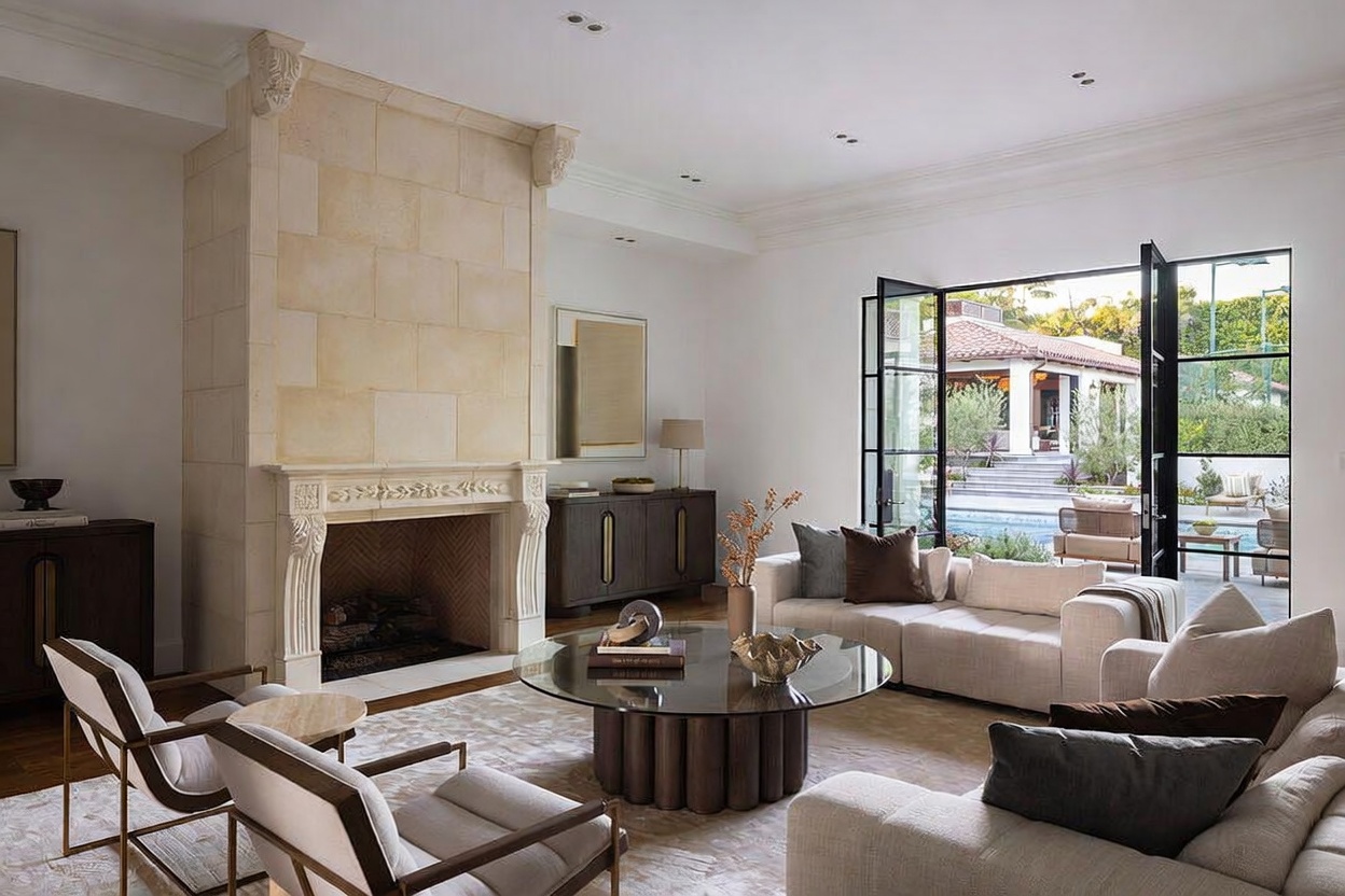 This elegant living room features a prominent stone-clad chimney breast with intricate carvings above the fireplace, serving as the room's focal point. The space is furnished with a neutral-toned sectional sofa, a circular glass-topped coffee table, and two modern armchairs, all arranged on a textured area rug. Large glass doors open to reveal a view of a swimming pool and a landscaped outdoor area, creating a seamless transition between indoor and outdoor living spaces.