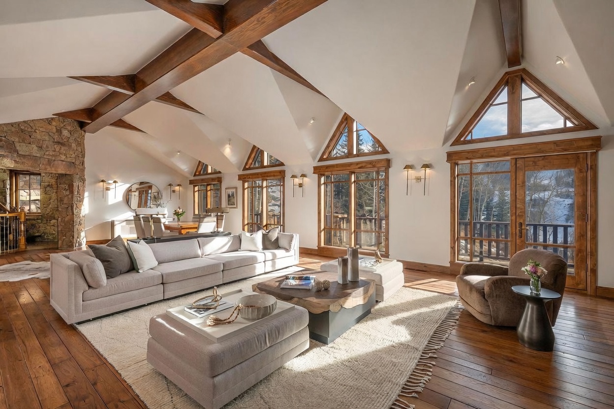 This expansive living room features vaulted ceilings with exposed timber beams and a prominent stone-clad chimney breast. The space is anchored by a large, neutral-toned sectional sofa and a unique wood-slab coffee table, all set upon a textured area rug. Large windows and glass doors provide abundant natural light and views of the surrounding landscape, creating an airy and sophisticated mountain-inspired atmosphere.