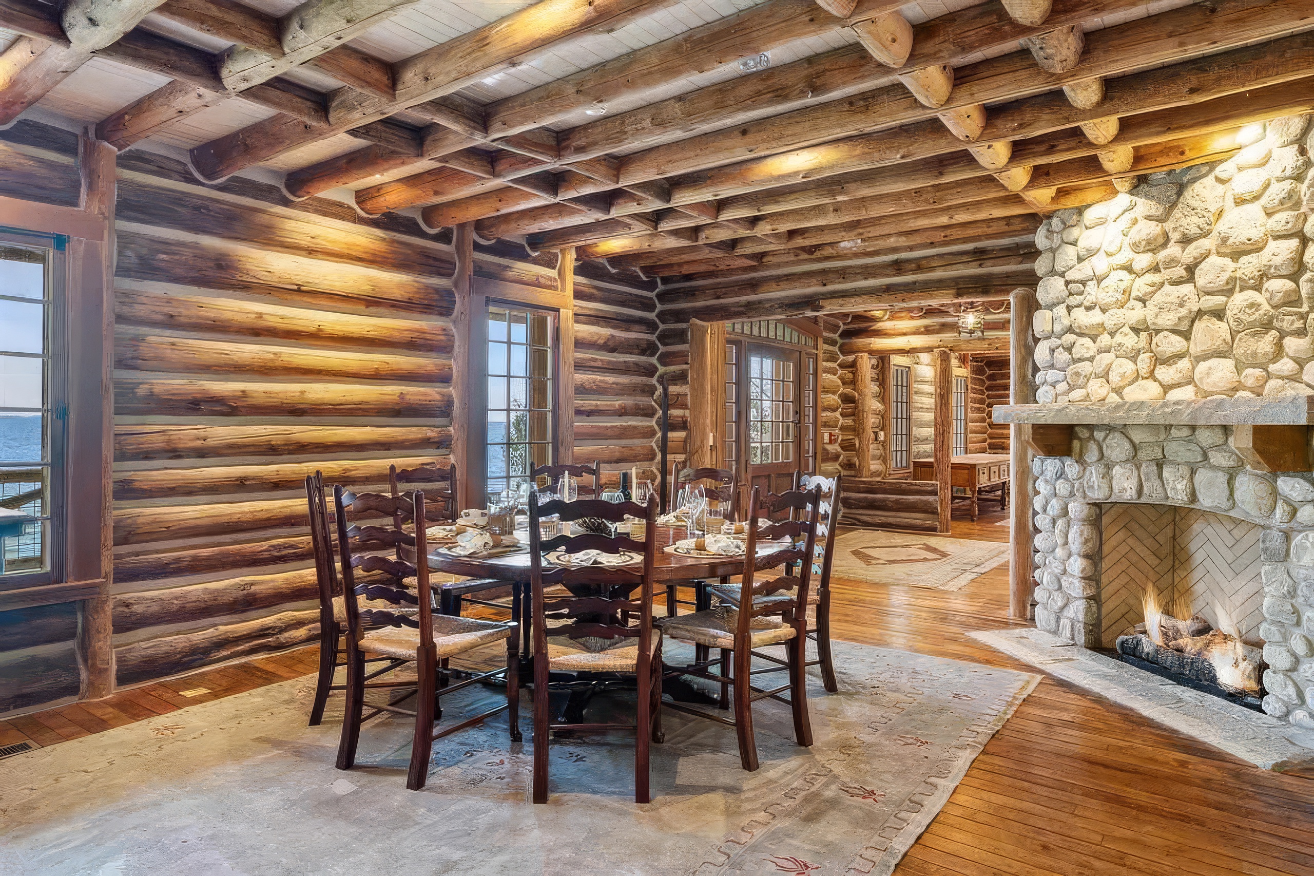 This rustic dining room features exposed log walls and a heavy timber-beamed ceiling, creating a warm and authentic cabin atmosphere. A round wooden dining table with six matching chairs sits atop a large, patterned area rug, positioned near a stone-clad chimney breast with a burning fire. The space is illuminated by natural light from large windows that offer a view of the water, enhancing the cozy and inviting aesthetic.