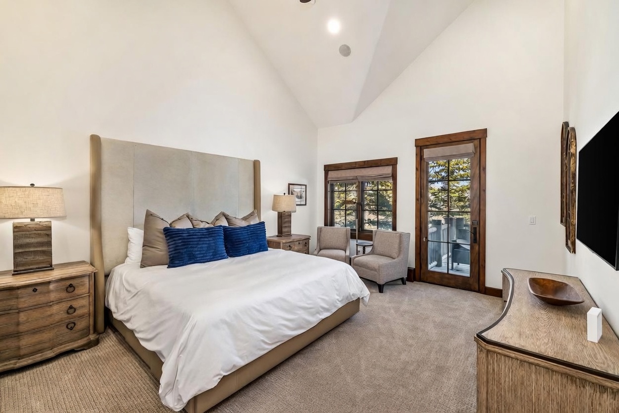 This spacious primary bedroom features a high vaulted ceiling and a large, neutral-toned upholstered headboard as the focal point. The room includes a comfortable seating area near a glass door that opens to an outdoor deck, providing ample natural light. A wooden dresser and matching nightstands add a warm, rustic touch to the clean, contemporary aesthetic.