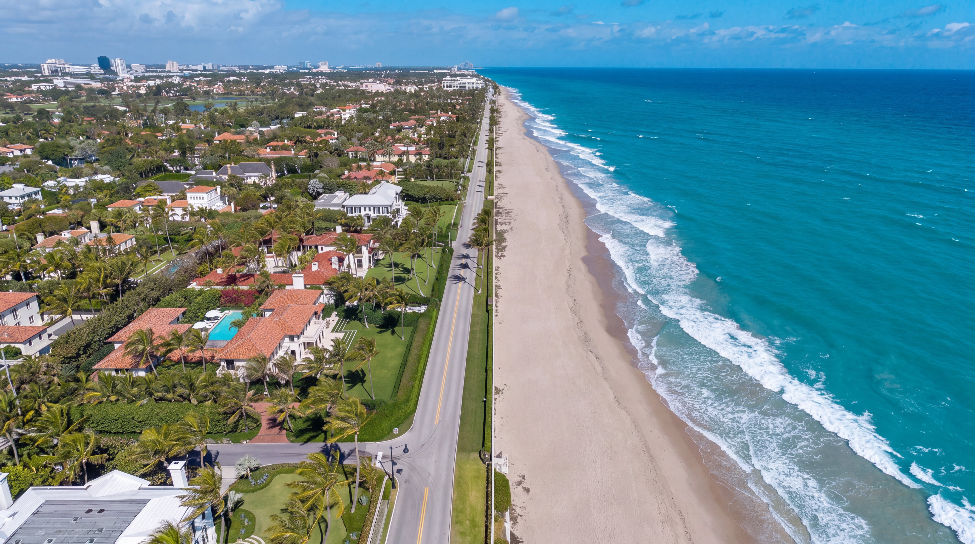 This high-altitude aerial view captures a luxurious beachfront estate situated between a coastal road and the Atlantic Ocean. The property features a sprawling residence with a terracotta-tiled roof, a private swimming pool, and lush landscaping, all highlighted by a white boundary outline. The perspective emphasizes the prime location, showcasing the expansive sandy beach and the vibrant turquoise water stretching toward the horizon.