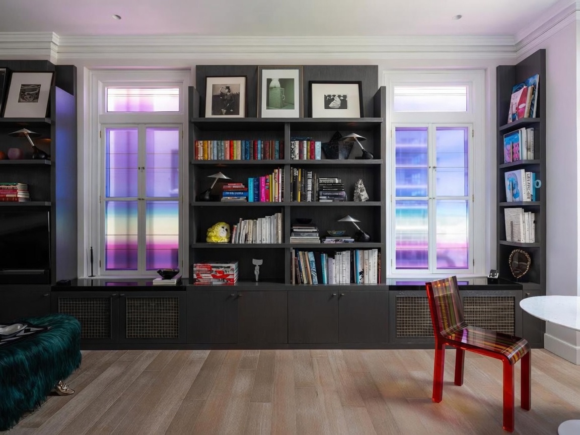 This sophisticated living space features a large, dark-toned custom built-in shelving unit that spans the wall, housing an extensive book collection and decorative art. Two windows with vibrant, multi-colored gradient film filters flank the central shelving, casting a unique light into the room. A modern, translucent red chair sits on the light wood flooring, adding a bold pop of color to the otherwise moody and curated aesthetic.