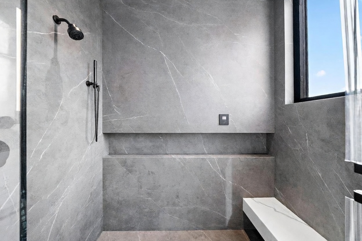 This modern walk-in shower features large-format gray stone tiles with subtle white veining throughout the walls. The space includes a built-in stone bench, a recessed wall niche for storage, and a sleek black shower fixture set. A window to the right allows natural light to brighten the minimalist, high-end design.